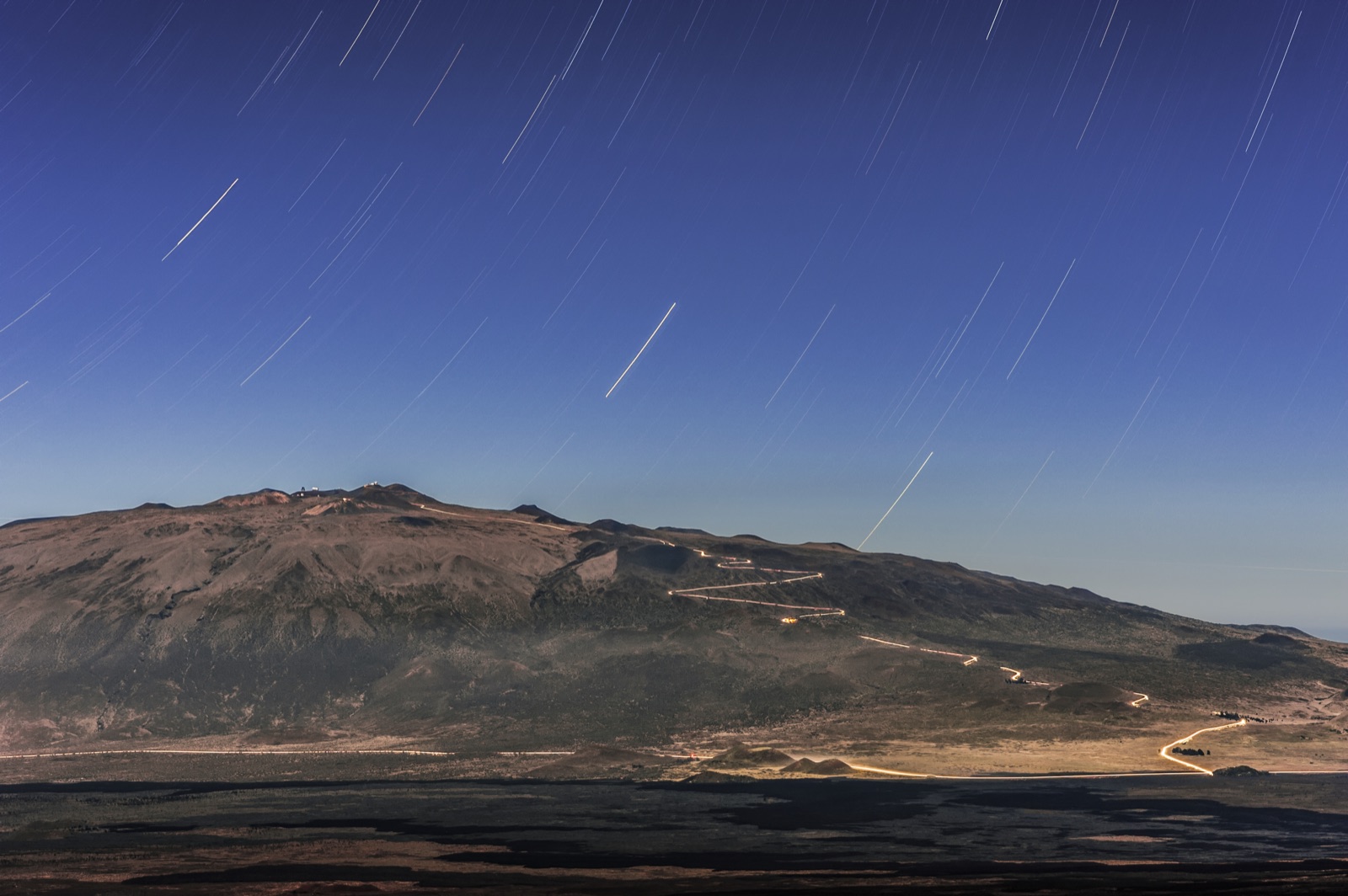 Star trails wheeling over the moonlit bulk of Maunakea, with the summit road and observatory lights visible.