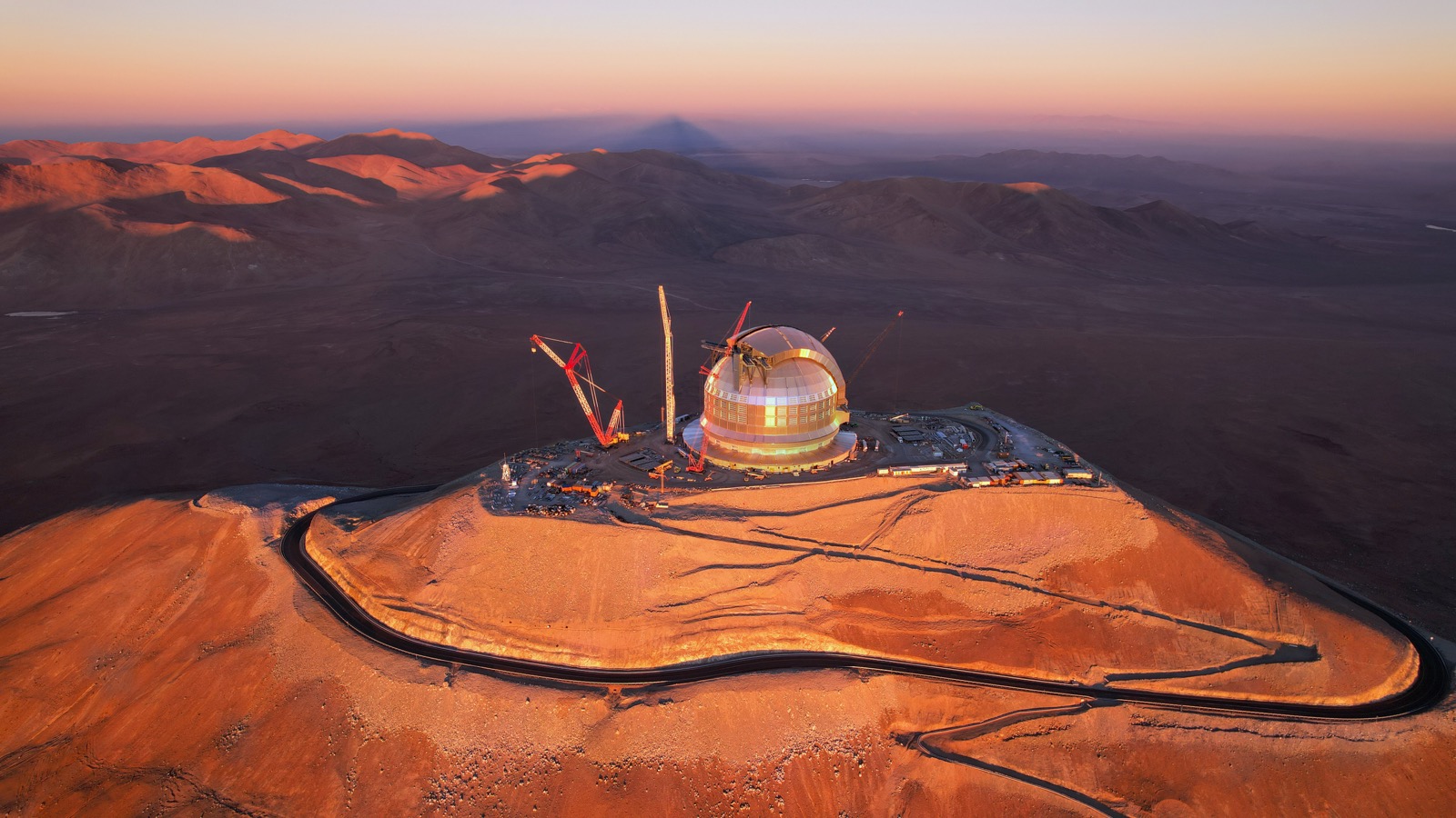 The Extremely Large Telescope's dome under construction on Cerro Armazones at sunset, surrounded by cranes.